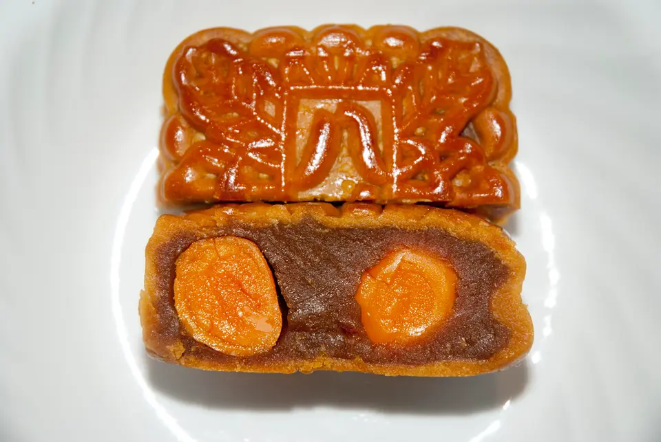Kue bulan yang dipotong melintang memperlihatkan dua kuning telur bebek asin utuh dalam pasta teratai gelap, dengan satu kue utuh di belakangnya