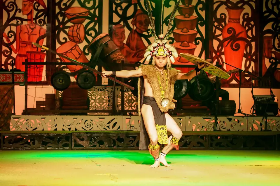 彫りを施した舞台で、盾とサイチョウの羽根の被り物を身につけ、イバンの戦の踊りンガジャットを舞う踊り手