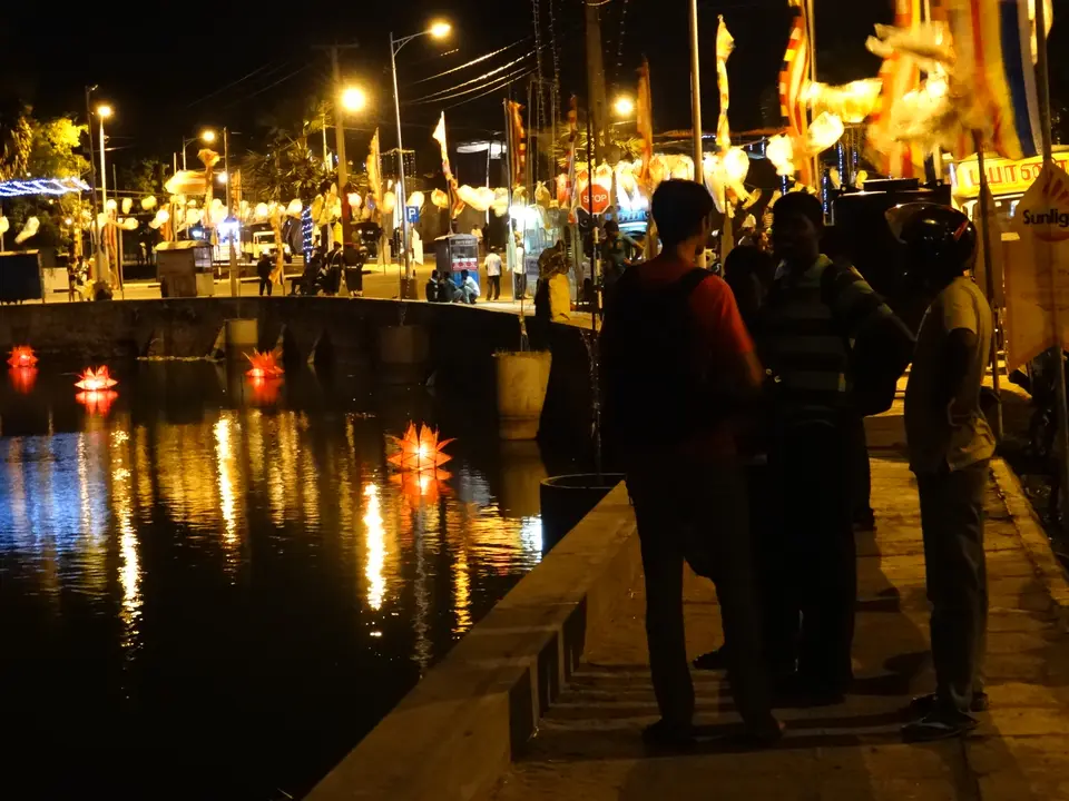 Vesak-Laternen treiben nachts auf dem Wasser in Jaffna, ihre Spiegelungen ziehen sich über die Fläche, während Menschen vom Ufer zusehen