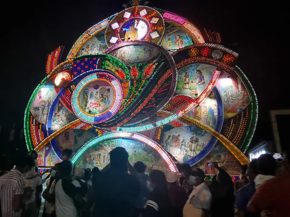 Ein riesiges beleuchtetes Vesak-Pandal in Maharagama bei Nacht, dessen bemalte Tafeln über einer Menge aus Schatten eine Jataka-Geschichte erzählen
